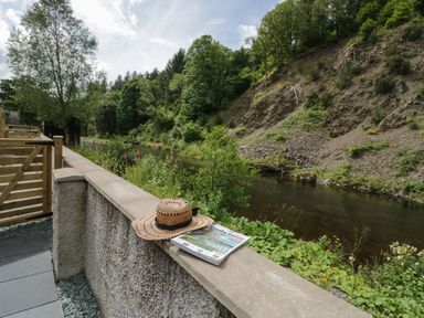 An outdoor patio with a wicker hat and magazine on a stone wall overlooking a river with trees and bushes at Riverside Cottage No 5 in Keswick