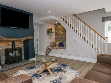 A living room with a glass coffee table on a round rug and a TV above a wood stove at Garden Cottage No 1 in Keswick