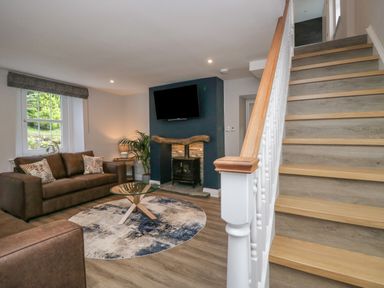 A living room with a brown sofa a round rug a glass coffee table a fireplace with a TV mounted above and stairs on the right side at Garden Cottage No 1 in Keswick
