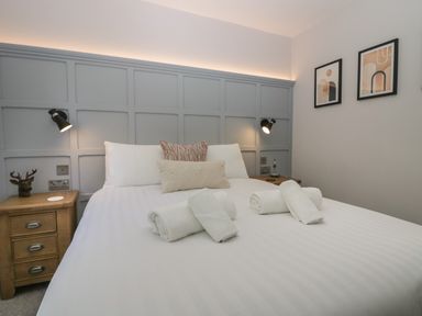 A bedroom with a large bed with white linens and rolled towels on it two wooden bedside tables with lamps and framed pictures on the wall at Garden Cottage No 1 in Keswick