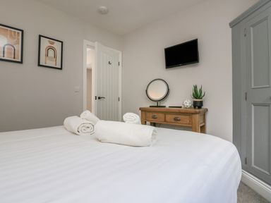 A bedroom with a bed and rolled towels a wooden desk with a round mirror and potted plant a wall mounted television and a grey wardrobe at Garden Cottage No 1 in Keswick
