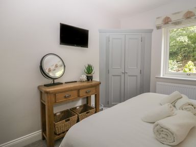 A bedroom with a wooden dresser a mirror a plant a wall mounted TV a window and a wardrobe at Garden Cottage No 1 in Keswick