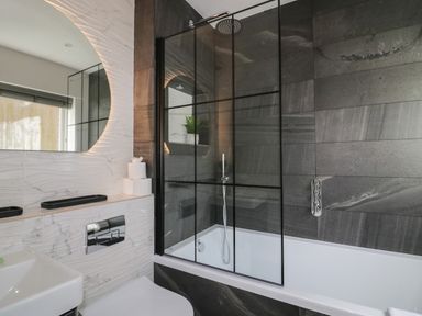 A bathroom with a bathtub shower enclosure a large round mirror and a stack of toilet paper at Garden Cottage No 1 in Keswick