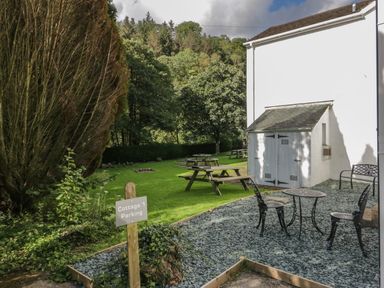 An outdoor area with picnic tables on grass and a small patio with metal chairs and table beside a white building at Garden Cottage No 1 in Keswick