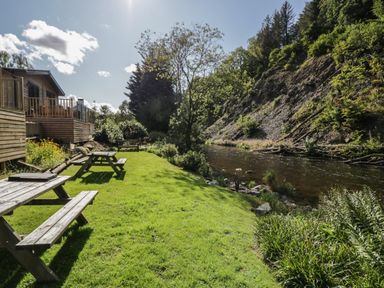 A grassy riverside area with wooden picnic tables benches and a cabin on the left at Garden Cottage No 1 in Keswick