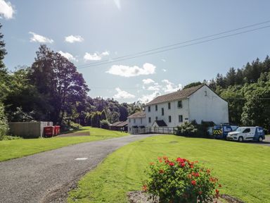 A two-story white building next to a driveway with parked vans and a flower bed at Garden Cottage No 1 in Keswick