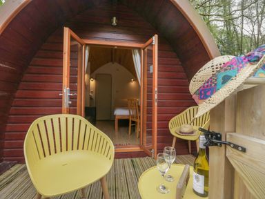 A wooden pod with open doors leading to a bedroom a small deck with two yellow chairs a table with two wine glasses a bottle of wine and a straw hat with colorful trim at Pod No 5 in Keswick