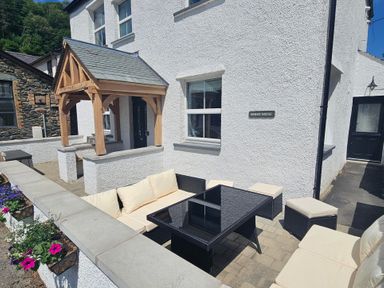 An outdoor patio with cushioned seating and a black glass table in front of a white house with a wooden porch at Briery House in Keswick