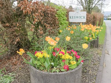 A roadside flower planter with yellow and red flowers near a welcome to Keswick sign at Briery House in Keswick