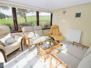 A sitting room with armchairs sofas a glass coffee table and large windows showing a green outdoor area at Pinegarth in Catton near Allendale