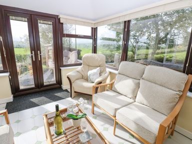 A sitting area with a sofa a chair and a glass table with a wine bottle and glasses in a room with large windows at Pinegarth in Catton near Allendale