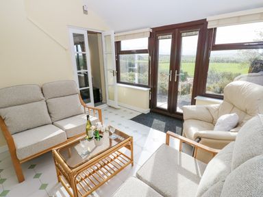 A sitting area with cushioned chairs a glass coffee table and large windows overlooking a green landscape at Pinegarth in Catton near Allendale