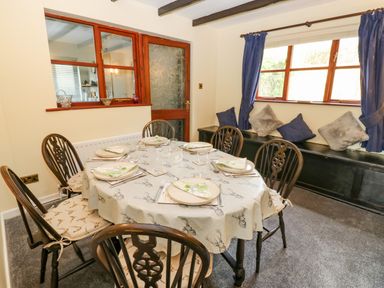 A dining room with a round table set for six people and wooden chairs near a window with blue curtains at Pinegarth in Catton near Allendale