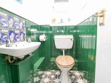 A bathroom with a white toilet with a wooden seat a small sink with faucets green panelled walls and patterned floor tiles at Pinegarth Catton near Allendale