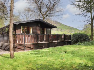 A wooden cabin with a fenced porch surrounded by grass and trees at Derwent Lodge in Keswick