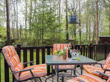 An outdoor wooden deck with a glass table and four striped cushioned chairs surrounded by trees at Derwent Lodge in Keswick