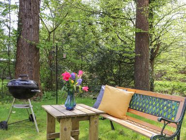 A wooden bench with pillows next to a wooden table with a vase of flowers and a black charcoal grill in a garden at Derwent Lodge in Keswick