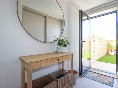 A wooden console table with plants under a round mirror next to an open door leading to a fenced garden at 11 Ebbtide in Porth