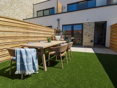 An outdoor seating area with a wooden dining table surrounded by wicker chairs on artificial grass at 11 Ebbtide in Porth