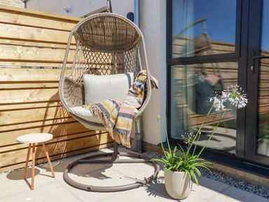 A hanging wicker chair with cushions and a blanket on a patio near a small round table and a potted plant at 11 Ebbtide in Porth