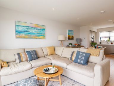A living room with a beige sectional sofa and pillows and paintings on the wall at 11 Ebbtide in Porth