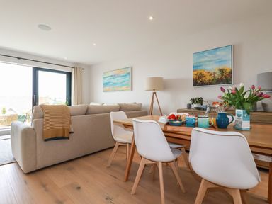 A dining area with four white chairs around a wooden table with flowers and kitchenware in a living room with a beige sofa and paintings on walls at 11 Ebbtide in Porth