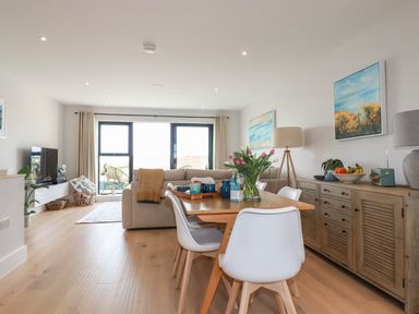 A dining table with white chairs and flowers next to a wooden sideboard and a living area with sofa and a TV at 11 Ebbtide in Porth