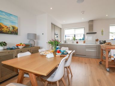 A kitchen and dining area with a wooden table and white chairs a sideboard with fruit and a lamp and kitchen cabinets with appliances at 11 Ebbtide in Porth