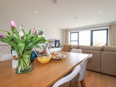 A dining table with flowers orange juice biscuits and chairs next to a sofa and large windows at 11 Ebbtide in Porth