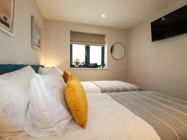 A bedroom with two beds white bedding yellow pillows a window with plants and a round mirror on the wall at 11 Ebbtide in Porth