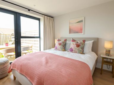 A bedroom with a bed covered with a pink blanket and patterned cushions next to a patio door at 11 Ebbtide in Porth