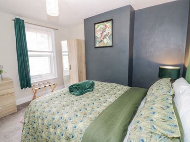 A bedroom with a large bed covered in peacock feather patterned bedding a wooden wardrobe a chest of drawers and a window with green curtains at Dobbies Den in Bewdley