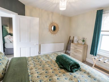 A bedroom with a bed covered with a peacock feather patterned duvet a wooden chest of drawers a round mirror on the wall and a window with curtains at Dobbies Den in Bewdley