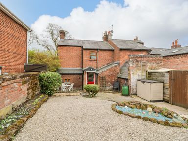 A backyard with gravel, a small blue rock garden, bushes, a brick house with a red door and outdoor seating at Dobbies Den in Bewdley