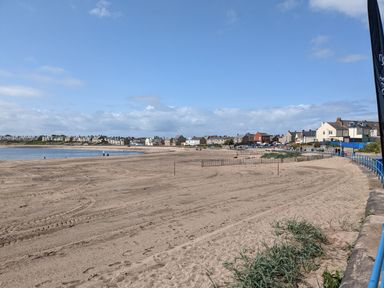 A sandy beach with footprints and grass near a blue railing with houses in the background at Fairwinds in Newbiggin-By-The-Sea