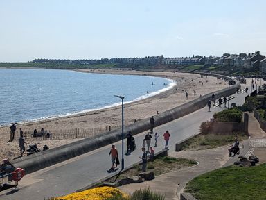 A beach with people walking on a promenade and houses along the shore at Fairwinds in Newbiggin-By-The-Sea