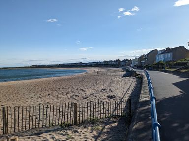 A sandy beach with a curved shoreline and a pathway with blue railings next to houses at Fairwinds in Newbiggin-By-The-Sea