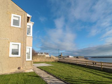 A grassy yard with a concrete path next to a building overlooking a fenced beach and ocean at Fairwinds in Newbiggin-By-The-Sea