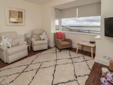 A sitting area with two beige armchairs a brown armchair a wooden side table a window with a sea view and a wall-mounted television at Fairwinds in Newbiggin-By-The-Sea