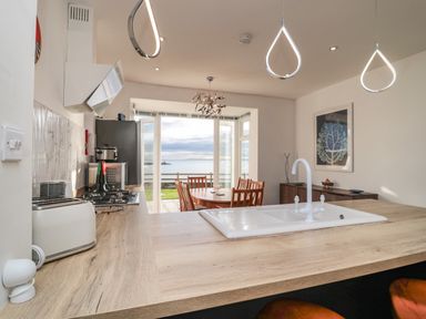 A kitchen with a countertop sink and wood dining table with chairs near a glass door overlooking the sea at Fairwinds in Newbiggin-By-The-Sea