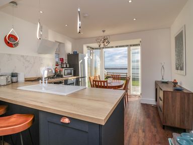 A kitchen with a wooden countertop and white sink opening into a dining area with a round table and wooden chairs overlooking a waterfront at Fairwinds in Newbiggin-By-The-Sea