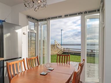 A dining area with a wooden table and chairs beside glass doors overlooking a garden and water at Fairwinds in Newbiggin-By-The-Sea