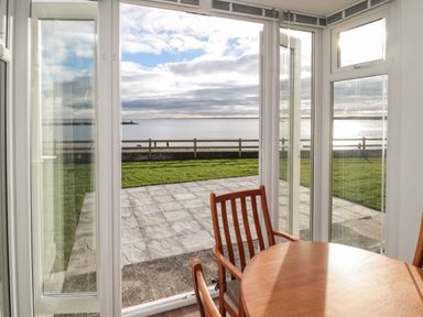 A dining area with wooden chairs and table near glass doors opening to a patio with grass and a sea view at Fairwinds in Newbiggin-By-The-Sea