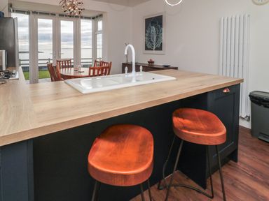 A kitchen island with a sink and two wooden stools with a dining table and chairs by windows overlooking the sea at Fairwinds in Newbiggin-By-The-Sea