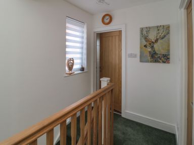 A hallway with a wooden banister a wooden door a window with blinds a clock above the door and a painting of a deer on the wall at Fairwinds in Newbiggin-By-The-Sea