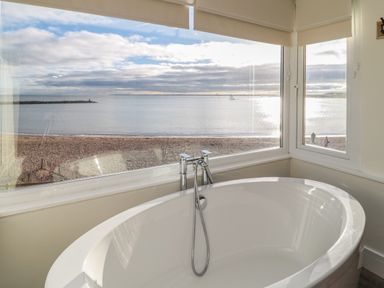 A bathroom with a white bathtub next to large windows overlooking a beach and sea at Fairwinds in Newbiggin-By-The-Sea