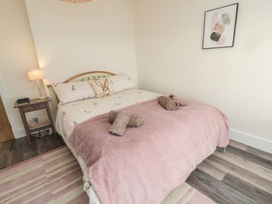 A bedroom with a double bed covered with a pink bedspread and rolled towels bedside table with a lamp and clock on a wooden floor at Fairwinds in Newbiggin-By-The-Sea