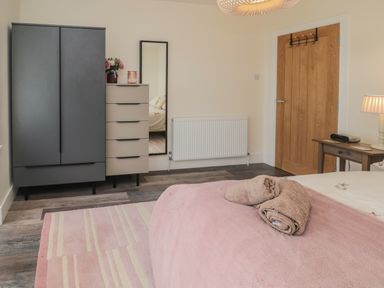 A bedroom with a bed covered by a pink blanket and towels on it a wooden door a radiator a dark wardrobe a chest of drawers with flowers and a mirror on the wall at Fairwinds in Newbiggin-By-The-Sea
