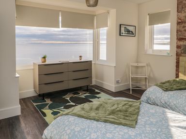 A bedroom with two beds a dresser a chair and windows overlooking the sea at Fairwinds in Newbiggin-By-The-Sea