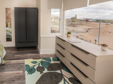 A bedroom with a black wardrobe a large beige dresser with plants on top and a window overlooking a beach at Fairwinds in Newbiggin-By-The-Sea
